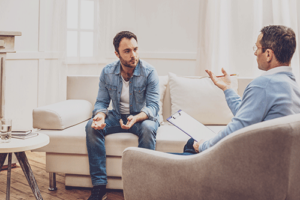 a man sits on a couch talking to his therapist nearby taking notes in the mans dual diagnosis rehab treatment 