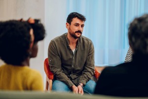 a man sits surrounded by group members in his heroin addiction rehab program and listens to what the other members are talking about