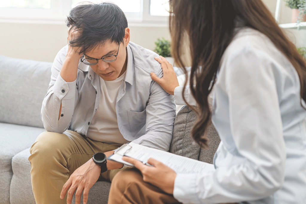 an upset man sits on the couch with his hand to his forehead while his therapist tells him the benefits of cognitive-behavioral therapy