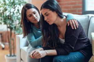 a woman hugs her friend while they sit on a couch and discuss the first step of heroin treatment