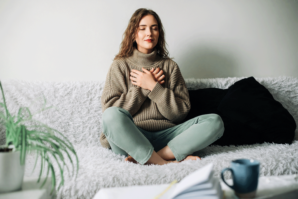 a young woman sits on her couch with her palms to her chest and meditating which is one of the coping skills for anxiety she learned