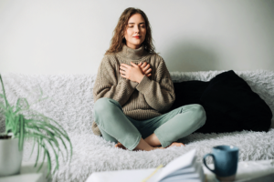 a young woman sits on her couch with her palms to her chest and meditating which is one of the coping skills for anxiety she learned